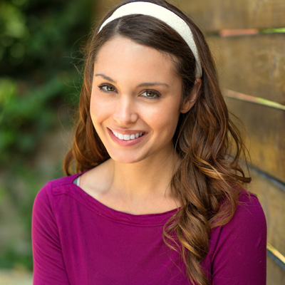 A woman with long hair, wearing a purple top and a headband, stands in front of a wooden fence.