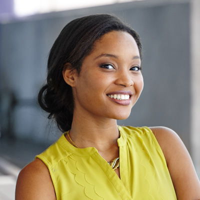 The image features a woman with a radiant smile, wearing a yellow top and standing against a backdrop of a building or structure.