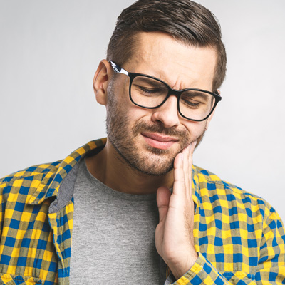 The image shows a man with glasses and a beard, wearing a yellow plaid shirt and holding his hand to his mouth. He appears to be in discomfort or pain, possibly due to toothache or a similar issue.